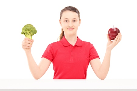 Smiling female teenager sitting and holding apple and broccoli isolated on white backgroundの写真素材