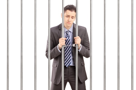 Handcuffed businessman in suit posing in jail and holding bars, isolated on white backgroundの写真素材