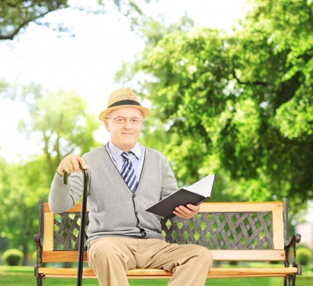 Senior man sitting on a wooden bench and reading a book, in a park, shot with a tilt and shift lensの写真素材