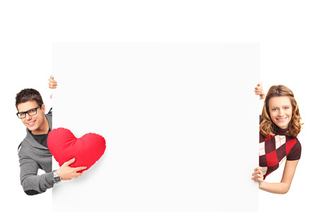 Smiling girl and boy with heart shaped pillow posing behind a white panel isolated on white backgroundの写真素材