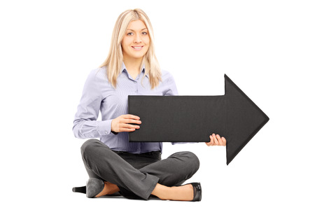 Young blond woman seated on floor holding a big black arrow pointing to the right isolated on white backgroundの写真素材
