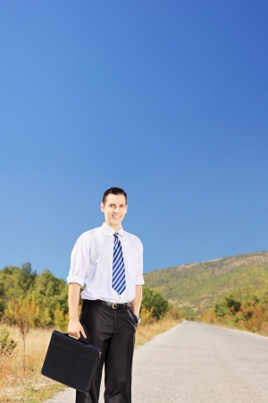 Young smiling businessperson holding a leather suitcase on an open roadの写真素材