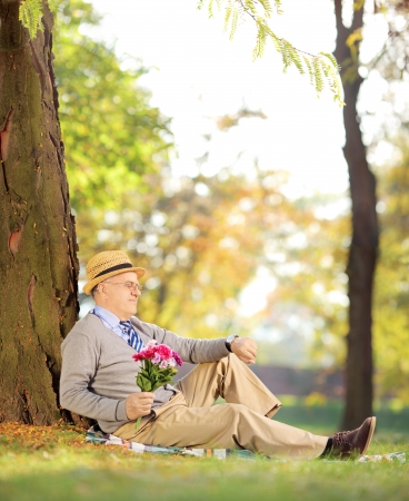 Senior gentleman with bunch of flowers sitting on a green grass and checking the time, in a park, shot with a tilt and shift lensの写真素材