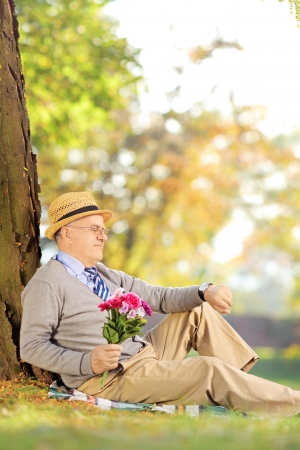 Senior gentleman with bunch of flowers sitting on a green grass and checking the time, in a parkの写真素材