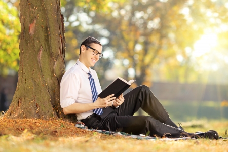 Young man sitting on a green grass and reading a book in park on a sunny dayの写真素材