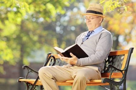 Senior man with hat sitting on a wooden bench and reading a book outsideの写真素材