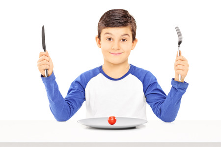 Young boy holding fork and knife with cherry tomato in front of him isolated on white backgroundの写真素材