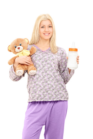 Young woman in pajamas holding teddy bear and a baby bottle isolated on white backgroundの写真素材