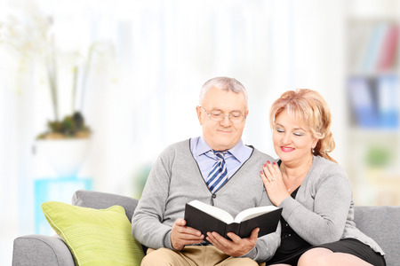 Mature couple reading a book seated on couch, at homeの写真素材