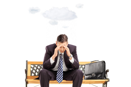 Stressed young businessman sitting on bench with cloud over head isolated on white の写真素材