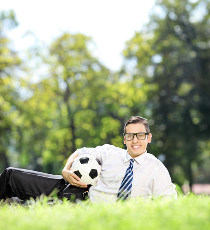 Young man lying on grass and holding a football in park shot with tilt and shift lensの写真素材
