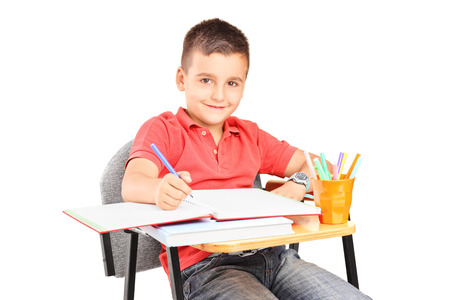 Schoolboy writing in a notebook seated on a school desk isolated on white backgroundの写真素材