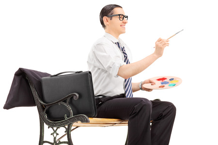 Businessman holding a paintbrush and a color palette seated on a wooden bench isolated on white backgroundの写真素材