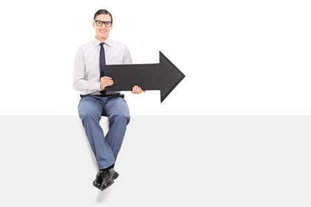 Cheerful man sitting on a panel and holding an arrow isolated on white background