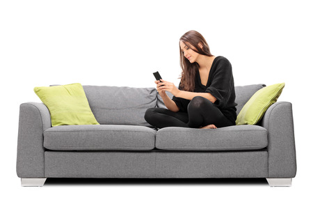 Young woman playing on her cell phone seated on a sofa isolated on white backgroundの写真素材