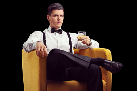 Serious man sitting in an armchair and drinking whiskey on black backgroundの写真素材