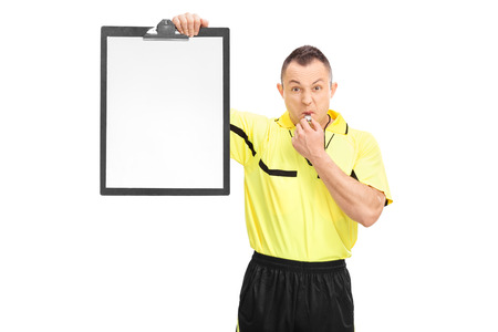 Angry football referee in a yellow shirt blowing a whistle and showing a blank paper on a clipboard isolated on white backgroundの写真素材