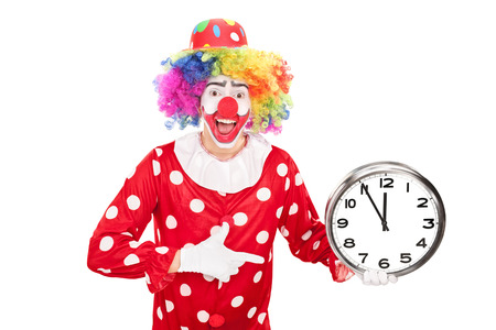 Young male clown holding a big wall clock in one hand and pointing towards the clock with the other isolated on white backgroundの写真素材