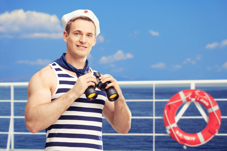 Young sailor holding binoculars and standing on a deck of a boat with blue sky and sea in the backgroundの写真素材