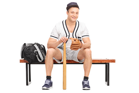 Young baseball player with a baseball glove and a bat sitting on bench and looking at the camera isolated on white backgroundの写真素材