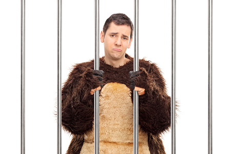 Studio shot of a sad young man in a bear costume standing behind bars in a cell isolated on white backgroundの写真素材