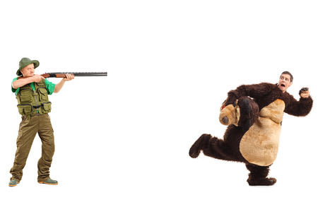 Full length portrait of a mature hunter aiming a rifle towards a man in a bear costume isolated on white backgroundの写真素材