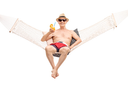 Carefree senior in red swim trunks lying in a hammock and drinking a delicious orange cocktail isolated on white backgroundの写真素材