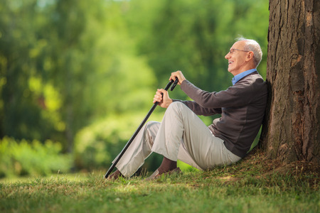 Senior gentleman sitting by a tree in a park and holding a cane on a beautiful summer dayの写真素材
