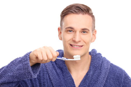 Young man in a blue bathrobe brushing his teeth with a toothbrush and looking at the camera isolated on white backgroundの写真素材