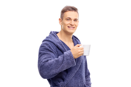 Young man in blue bathrobe holding a cup of coffee and looking at the camera isolated on white backgroundの写真素材