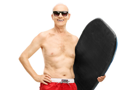 Senior man in blue swim trunks holding a surfboard and looking at the camera isolated on white backgroundの写真素材