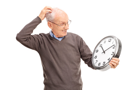 Confused senior holding a big wall clock and scratching his head isolated on white backgroundの写真素材