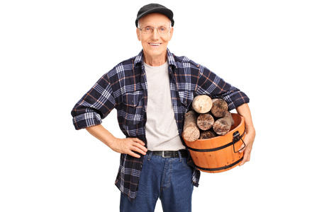 Cheerful senior in a blue checkered shirt holding a basket full of logs and smiling isolated on white backgroundの写真素材