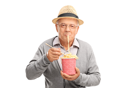 Joyful senior eating Chinese noodles from a paper box and looking at the camera isolated on white backgroundの写真素材