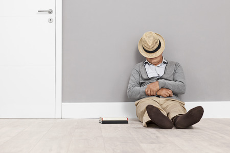 Senior sleeping on the floor with his hat covering his face and leaning against a wall next to a white doorの写真素材