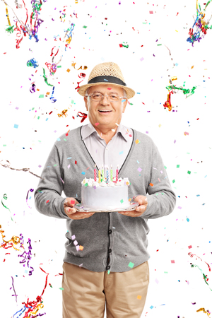 Vertical shot of a joyful senior gentleman carrying a birthday cake with party streamers flying all around him isolated on white backgroundの写真素材