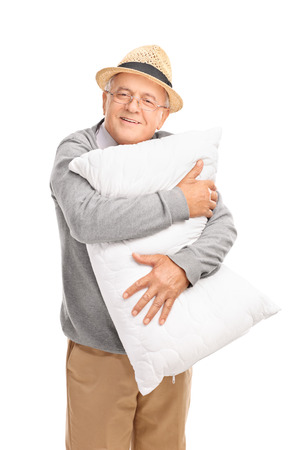 Vertical shot of a cheerful senior gentleman hugging a white pillow and looking at the camera isolated on white backgroundの写真素材