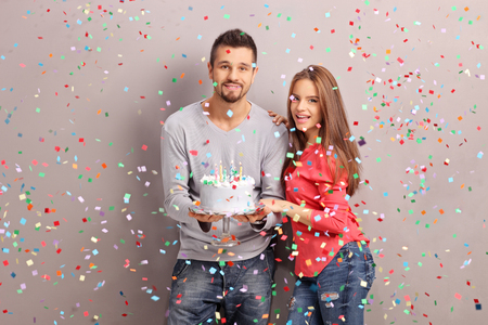 Young joyful couple holding a birthday cake with a lot of confetti streamers flying around themの写真素材