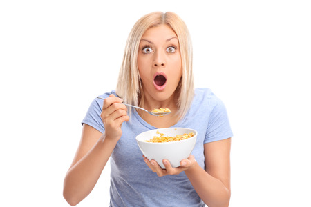 Shocked woman eating cereal from a bowl and looking at the camera isolated on white backgroundの写真素材