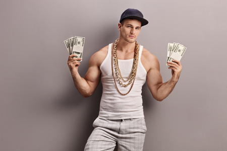 Young male rapper with golden chain around his neck holding few stacks of money and looking at the cameraの写真素材