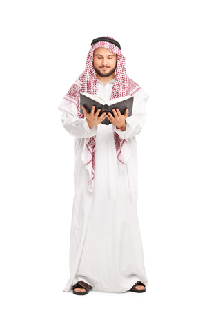Full length portrait of a young male Arab in a white robe and a checkered veil reading a book isolated on white backgroundの写真素材