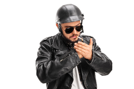 Studio shot of a male biker in a black leather jacket lighting up a cigarette with a lighter isolated on white backgroundの写真素材