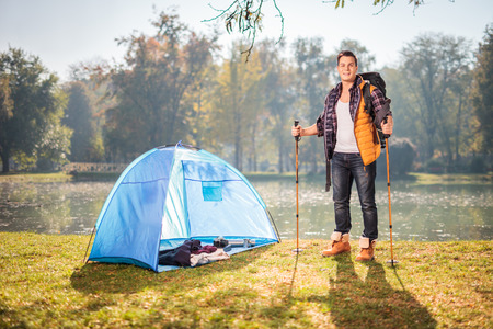 Young male hiker posing by a pond next to a blue tent in a field on a sunny autumn dayの写真素材