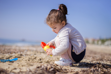 Adorable little girl playing with the sand on a beach with a little red plastic spade and a yellow cupの写真素材