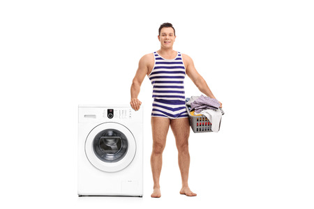 Young man in a striped underwear holding a laundry basket and standing next to a washing machine isolated on white backgroundの写真素材