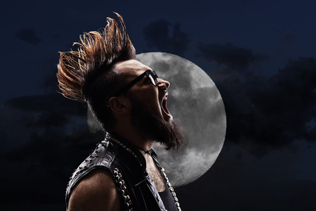 Young man with a Mohawk hairstyle shouting in the night in front of a full moonの写真素材