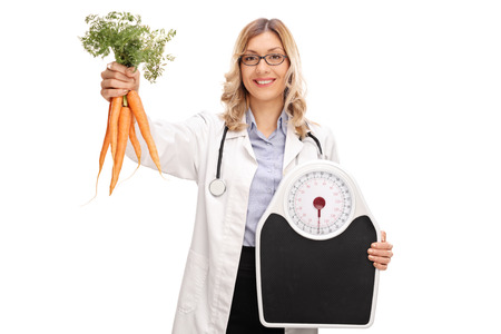 Cheerful female doctor holding a bunch of carrots and a weight scale isolated on white backgroundの写真素材