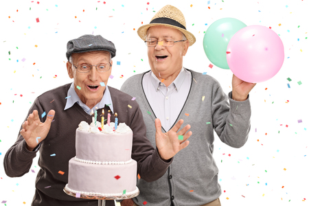 Two excited senior gentlemen blowing candles on a birthday cake isolated on white backgroundの写真素材