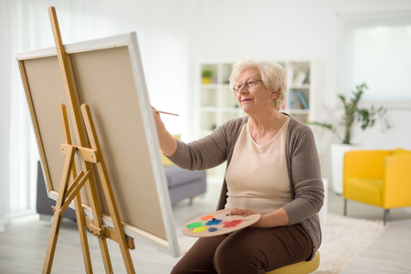 Mature lady painting on a canvas with a paintbrush at homeの写真素材
