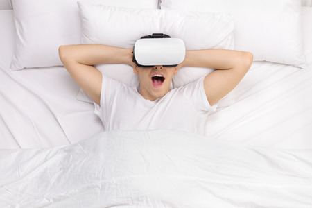 Joyful guy laying on a comfortable bed and watching something with VR goggles
